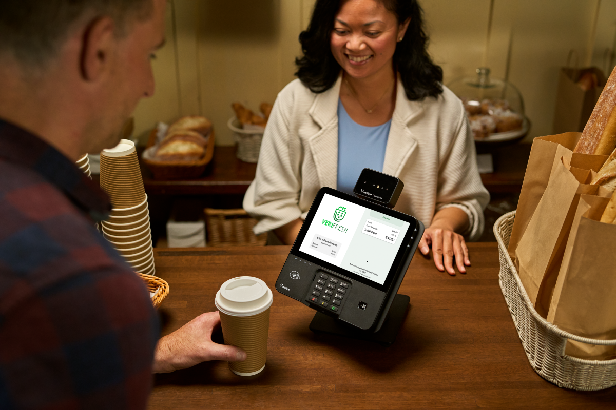 Customer holding a coffee cup at a bakery counter while a smiling cashier stands behind, with a digital payment terminal displaying a Verifresh transaction.